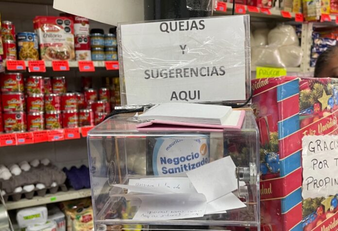 Escuchar para vender Caja de sugerencias y quejas colocada en el mostrador de una frutería, rodeada de productos, como muestra de comunicación directa entre comerciantes y clientes.