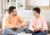¿Hablas con tus hijos? Padre e hijo adolescentes sentados en una sala de estar, conversando de manera cercana y relajada, generando un ambiente de confianza.
