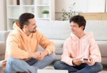 ¿Hablas con tus hijos? Padre e hijo adolescentes sentados en una sala de estar, conversando de manera cercana y relajada, generando un ambiente de confianza.