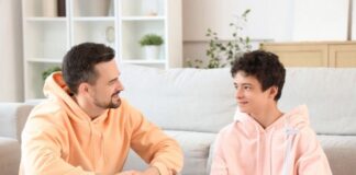 ¿Hablas con tus hijos? Padre e hijo adolescentes sentados en una sala de estar, conversando de manera cercana y relajada, generando un ambiente de confianza.