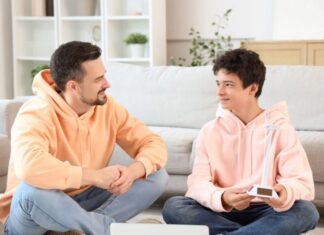 ¿Hablas con tus hijos? Padre e hijo adolescentes sentados en una sala de estar, conversando de manera cercana y relajada, generando un ambiente de confianza.