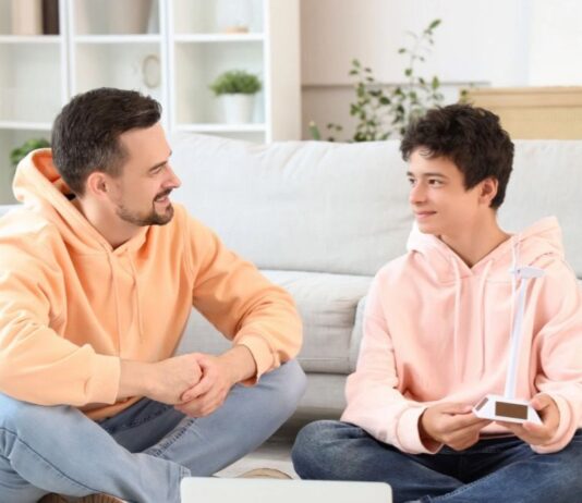 ¿Hablas con tus hijos? Padre e hijo adolescentes sentados en una sala de estar, conversando de manera cercana y relajada, generando un ambiente de confianza.