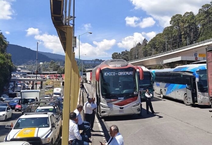 Bloqueo de autobuses y camiones en carretera con operadores detenidos al costado de la vía, generando tráfico vehicular.