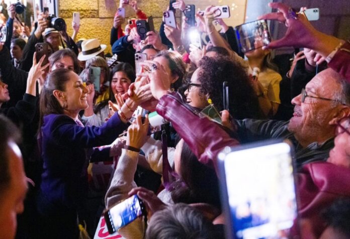 Presidenta Claudia Sheinbaum saludando a una multitud de personas que la reciben con celulares y aplausos en Barcelona.