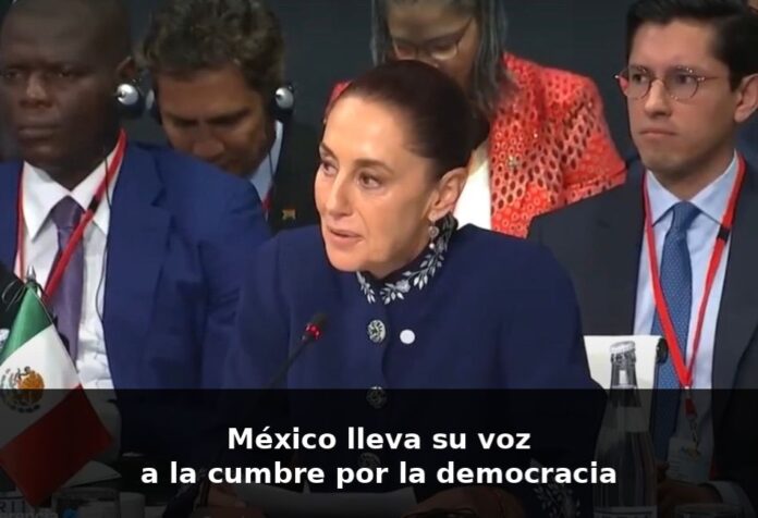 Claudia Sheinbaum habla en un foro internacional en Barcelona, sentada frente a un micrófono, con la bandera de México visible a un costado.