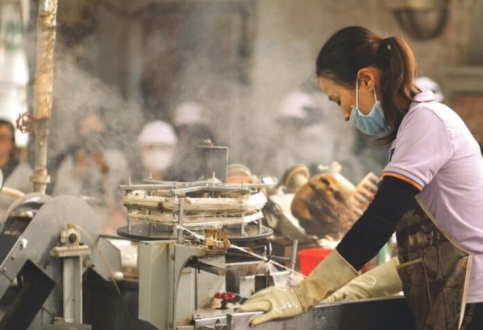 Bajas tasas de desempleo Mujer trabajando en una línea de producción industrial mientras opera maquinaria en un entorno de manufactura, con vapor y equipo de trabajo al fondo.