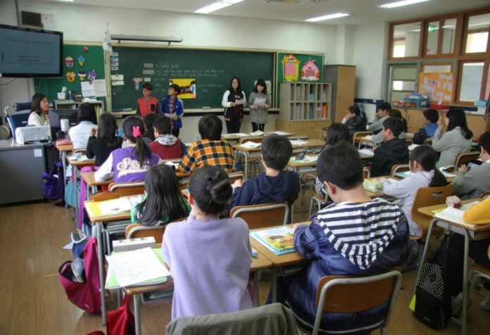Alumnos de educación básica en un salón de clases mexicano atienden una exposición frente al pizarrón, donde se observan materiales educativos, mensajes de convivencia escolar y la bandera nacional.