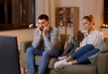 ¿Por qué cada vez vemos más sexo y drogas en películas y series? Familia sentada frente al televisor durante la noche mientras observa contenido audiovisual en un ambiente doméstico iluminado por la luz de la pantalla.