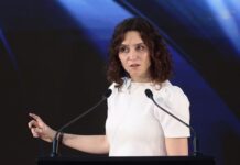 Ayuso regresa a España y describe un México que millones de mexicanos no reconocen Isabel Díaz Ayuso durante una conferencia en México, frente a dos micrófonos y con fondo oscuro iluminado en tonos azules.