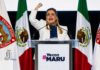 Maru Campos endurece su discurso contra Morena; el conflicto político entra en una nueva fase Maru Campos pronuncia un discurso desde un podio, con el puño levantado, frente a banderas de México y del estado de Chihuahua durante un acto político de apoyo.
