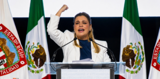 Maru Campos endurece su discurso contra Morena; el conflicto político entra en una nueva fase Maru Campos pronuncia un discurso desde un podio, con el puño levantado, frente a banderas de México y del estado de Chihuahua durante un acto político de apoyo.