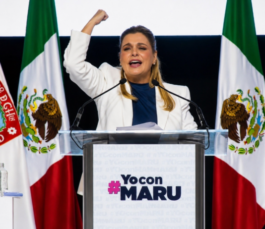 Maru Campos endurece su discurso contra Morena; el conflicto político entra en una nueva fase Maru Campos pronuncia un discurso desde un podio, con el puño levantado, frente a banderas de México y del estado de Chihuahua durante un acto político de apoyo.