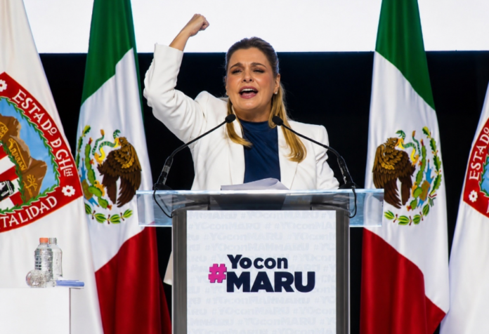 Maru acampos intensifica batalla Maru Campos pronuncia un discurso desde un podio, con el puño levantado, frente a banderas de México y del estado de Chihuahua durante un acto político de apoyo.