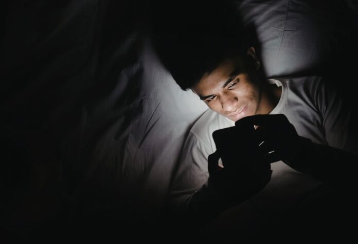 Persona acostada en una cama utilizando un teléfono celular en una habitación oscura durante la noche, iluminada únicamente por la luz de la pantalla.