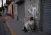 La indigencia: el rostro más extremo de la exclusión social Persona en situación de calle cubierta con una cobija, sentada junto a una pared y cortinas metálicas cerradas en una calle urbana al amanecer o atardecer.