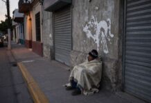 La indigencia: el rostro más extremo de la exclusión social Persona en situación de calle cubierta con una cobija, sentada junto a una pared y cortinas metálicas cerradas en una calle urbana al amanecer o atardecer.