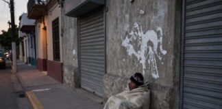 La indigencia: el rostro más extremo de la exclusión social Persona en situación de calle cubierta con una cobija, sentada junto a una pared y cortinas metálicas cerradas en una calle urbana al amanecer o atardecer.