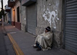 La indigencia: el rostro más extremo de la exclusión social Persona en situación de calle cubierta con una cobija, sentada junto a una pared y cortinas metálicas cerradas en una calle urbana al amanecer o atardecer.