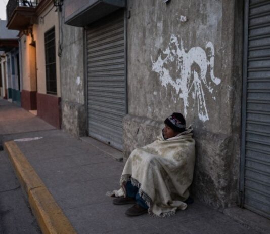 La indigencia: el rostro más extremo de la exclusión social Persona en situación de calle cubierta con una cobija, sentada junto a una pared y cortinas metálicas cerradas en una calle urbana al amanecer o atardecer.