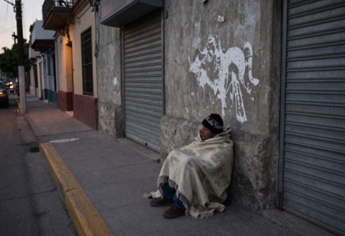 Persona en situación de calle cubierta con una cobija, sentada junto a una pared y cortinas metálicas cerradas en una calle urbana al amanecer o atardecer.