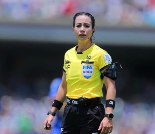 Katia Itzel García se prepara para otro capítulo histórico en el arbitraje mexicano Katia Itzel García, árbitra mexicana con uniforme amarillo de la FIFA, observa una jugada durante un partido en un estadio lleno de aficionados.