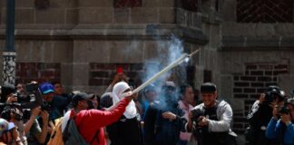 La CNTE inicia huelga nacional y reabre el debate sobre la educación de los niños Manifestantes participan en una protesta en el Zócalo de la Ciudad de México. Una persona sostiene un artefacto que emite humo mientras periodistas y fotógrafos documentan la movilización frente a un edificio histórico.