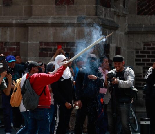 La CNTE inicia huelga nacional y reabre el debate sobre la educación de los niños Manifestantes participan en una protesta en el Zócalo de la Ciudad de México. Una persona sostiene un artefacto que emite humo mientras periodistas y fotógrafos documentan la movilización frente a un edificio histórico.
