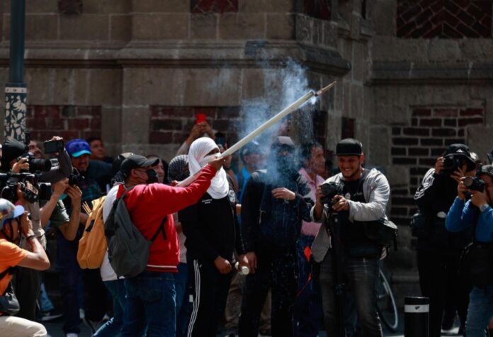 Manifestantes participan en una protesta en el Zócalo de la Ciudad de México. Una persona sostiene un artefacto que emite humo mientras periodistas y fotógrafos documentan la movilización frente a un edificio histórico.