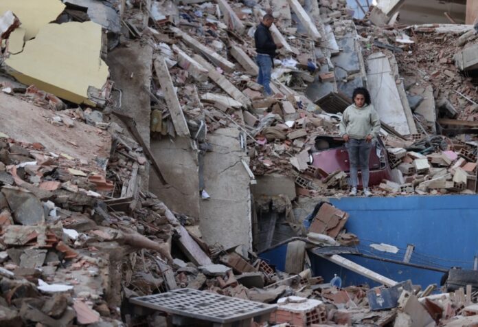 Pan de recuperación de Venezuela Mujer de pie entre los escombros de edificios derrumbados por los terremotos en Venezuela, con estructuras colapsadas y labores de recuperación en el lugar.