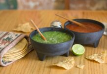 ¿La salsa quedó demasiado picosa? Todavía hay forma de suavizarla Dos molcajetes con salsa verde y salsa roja sobre una mesa de madera, acompañados de tortillas de maíz, totopos y un limón.