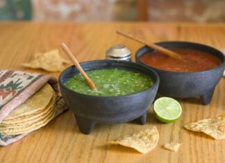 ¿La salsa quedó demasiado picosa? Todavía hay forma de suavizarla Dos molcajetes con salsa verde y salsa roja sobre una mesa de madera, acompañados de tortillas de maíz, totopos y un limón.