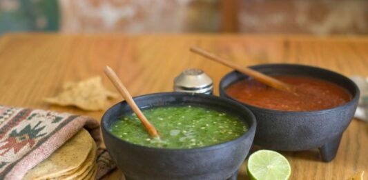 Dos molcajetes con salsa verde y salsa roja sobre una mesa de madera, acompañados de tortillas de maíz, totopos y un limón.
