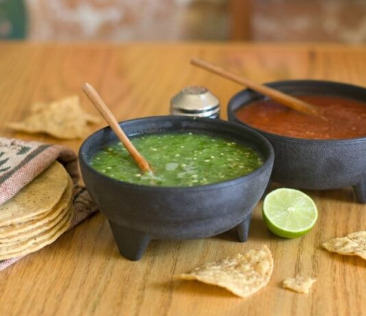 ¿La salsa quedó demasiado picosa? Todavía hay forma de suavizarla Dos molcajetes con salsa verde y salsa roja sobre una mesa de madera, acompañados de tortillas de maíz, totopos y un limón.