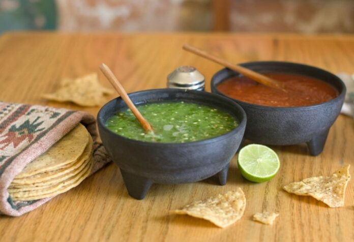 Dos molcajetes con salsa verde y salsa roja sobre una mesa de madera, acompañados de tortillas de maíz, totopos y un limón.