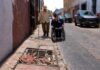 Las ciudades de México siguen poniendo obstáculos a adultos mayores y personas con discapacidad Adulto mayor con bastón camina por una banqueta estrecha y deteriorada, mientras una mujer en silla de ruedas circula por la calle junto a vehículos estacionados. En primer plano se observa un registro abierto con escombros que obstruye el paso peatonal.