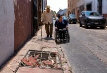Las ciudades de México siguen poniendo obstáculos a adultos mayores y personas con discapacidad Adulto mayor con bastón camina por una banqueta estrecha y deteriorada, mientras una mujer en silla de ruedas circula por la calle junto a vehículos estacionados. En primer plano se observa un registro abierto con escombros que obstruye el paso peatonal.