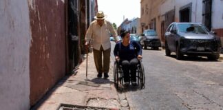 Las ciudades de México siguen poniendo obstáculos a adultos mayores y personas con discapacidad Adulto mayor con bastón camina por una banqueta estrecha y deteriorada, mientras una mujer en silla de ruedas circula por la calle junto a vehículos estacionados. En primer plano se observa un registro abierto con escombros que obstruye el paso peatonal.