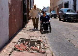 Las ciudades de México siguen poniendo obstáculos a adultos mayores y personas con discapacidad Adulto mayor con bastón camina por una banqueta estrecha y deteriorada, mientras una mujer en silla de ruedas circula por la calle junto a vehículos estacionados. En primer plano se observa un registro abierto con escombros que obstruye el paso peatonal.