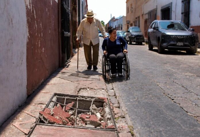 Adulto mayor con bastón camina por una banqueta estrecha y deteriorada, mientras una mujer en silla de ruedas circula por la calle junto a vehículos estacionados. En primer plano se observa un registro abierto con escombros que obstruye el paso peatonal.
