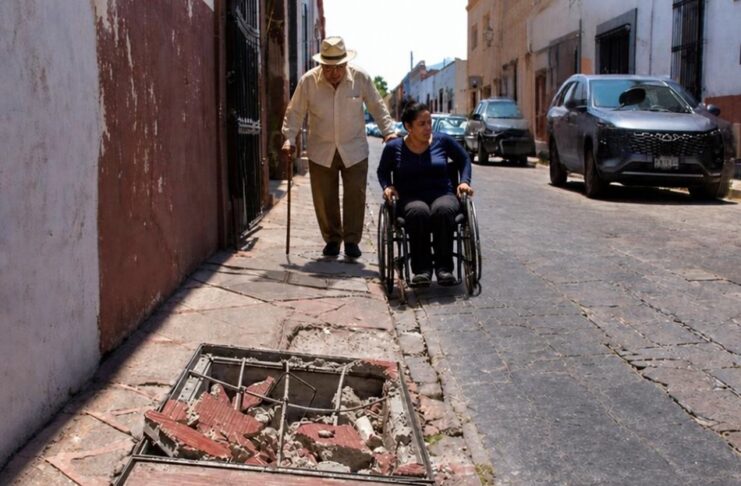 Las ciudades de México siguen poniendo obstáculos a adultos mayores y personas con discapacidad Adulto mayor con bastón camina por una banqueta estrecha y deteriorada, mientras una mujer en silla de ruedas circula por la calle junto a vehículos estacionados. En primer plano se observa un registro abierto con escombros que obstruye el paso peatonal.