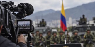 El efecto Bukele: la mano dura se convierte en espectáculo Imagen conceptual de una cámara de televisión enfocando un acto militar en Colombia, con soldados, vehículos y la bandera colombiana al fondo.