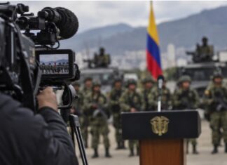 El efecto Bukele: la mano dura se convierte en espectáculo Imagen conceptual de una cámara de televisión enfocando un acto militar en Colombia, con soldados, vehículos y la bandera colombiana al fondo.