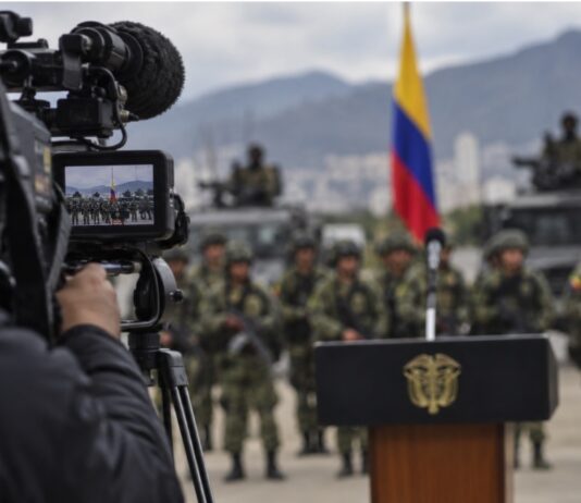 El efecto Bukele: la mano dura se convierte en espectáculo Imagen conceptual de una cámara de televisión enfocando un acto militar en Colombia, con soldados, vehículos y la bandera colombiana al fondo.