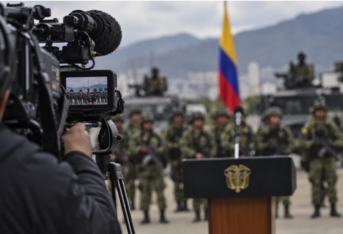 De la Espriella, efecto Bukele Imagen conceptual de una cámara de televisión enfocando un acto militar en Colombia, con soldados, vehículos y la bandera colombiana al fondo.