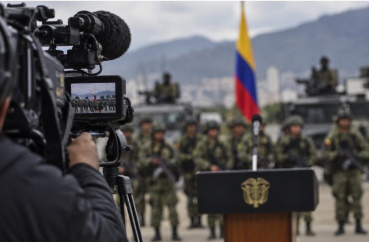 El efecto Bukele: la mano dura se convierte en espectáculo Imagen conceptual de una cámara de televisión enfocando un acto militar en Colombia, con soldados, vehículos y la bandera colombiana al fondo.