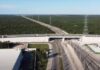 El sureste mexicano construye una nueva infraestructura para su desarrollo económico Vista aérea de una vía ferroviaria que pasa por debajo de un puente carretero, rodeada de extensas áreas de vegetación y acompañada por torres de transmisión eléctrica en el sureste de México.