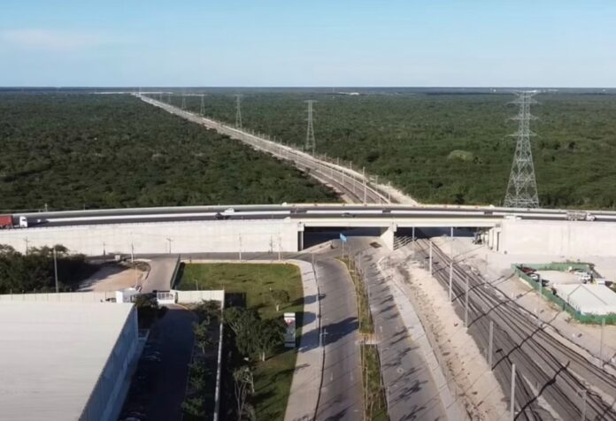 Infraestructura en el Sureste de México Vista aérea de una vía ferroviaria que pasa por debajo de un puente carretero, rodeada de extensas áreas de vegetación y acompañada por torres de transmisión eléctrica en el sureste de México.