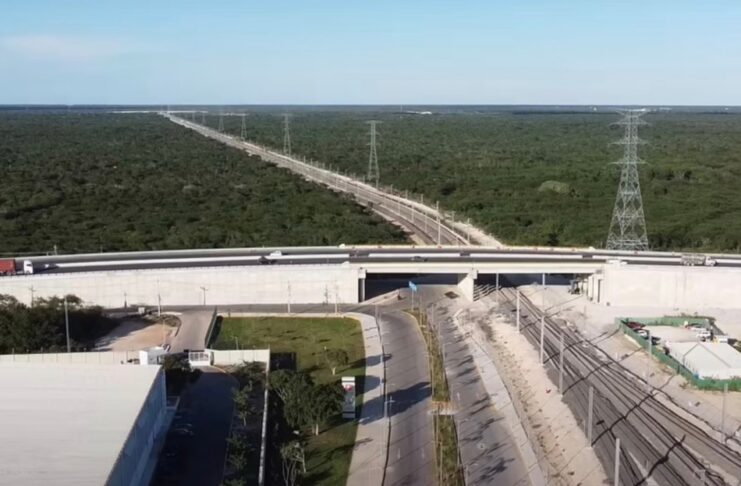 El sureste mexicano construye una nueva infraestructura para su desarrollo económico Vista aérea de una vía ferroviaria que pasa por debajo de un puente carretero, rodeada de extensas áreas de vegetación y acompañada por torres de transmisión eléctrica en el sureste de México.