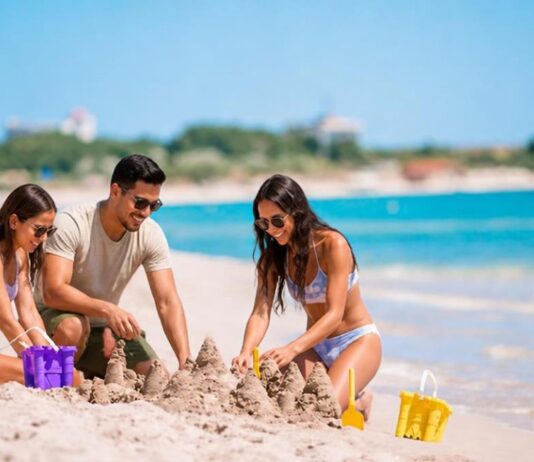Turismo en temporada baja: viajar mejor, gastar menos y descubrir más Tres personas construyen castillos de arena en una playa de aguas turquesas, con pocos visitantes alrededor. Dos mujeres y un hombre disfrutan de una actividad recreativa junto al mar.