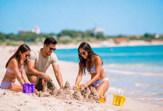 Temporada baja Tres personas construyen castillos de arena en una playa de aguas turquesas, con pocos visitantes alrededor. Dos mujeres y un hombre disfrutan de una actividad recreativa junto al mar.